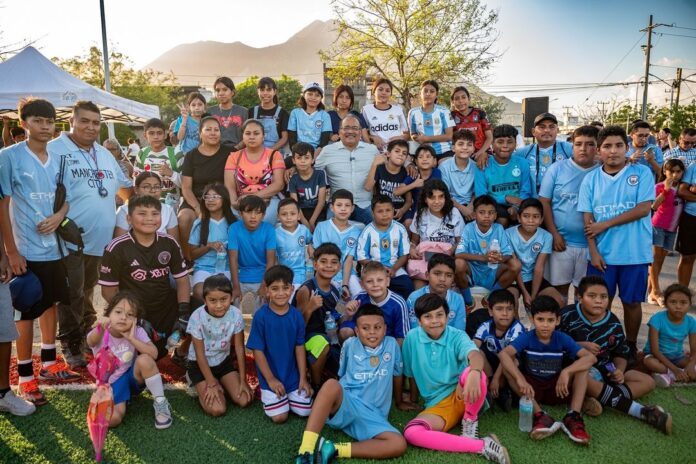 El Alcalde Andrés Mijes da la patada inicial en la nueva cancha de futbol rápido en la Colonia Privadas de Camino Real, acompañado por niños y jóvenes del área.